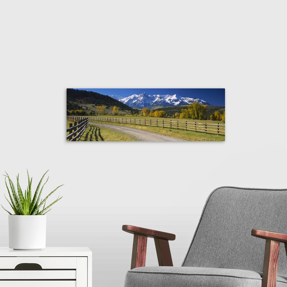 A modern room featuring Panoramic photograph of dirt path lined by a wooden gate with snow covered mountains in the dista...