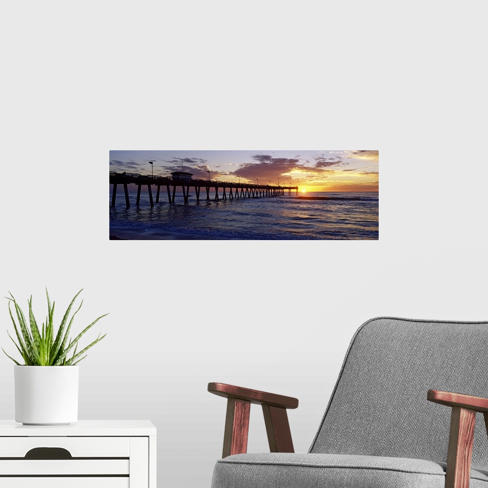 A modern room featuring A pier that reaches far out into the ocean is photographed from the beach with the sun just setti...