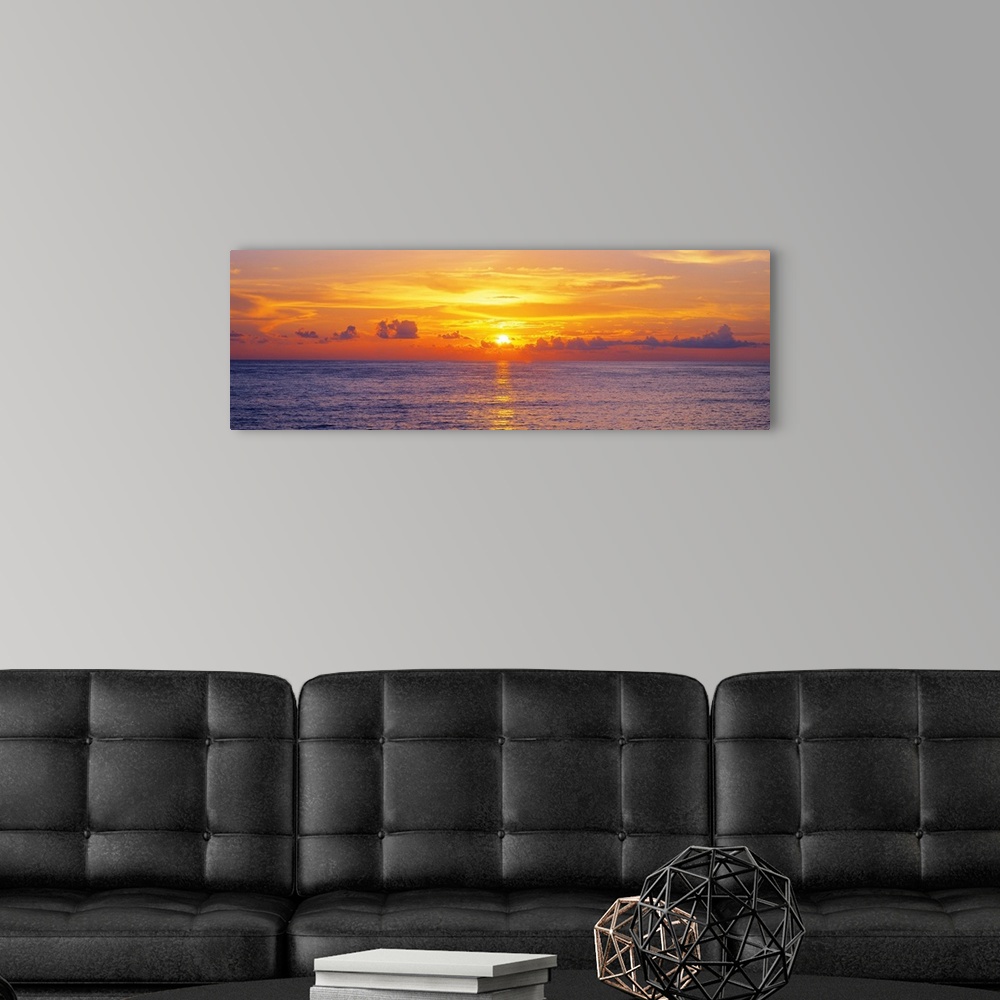A modern room featuring This panoramic seascape shows the sun setting behind clouds on the horizon in this photographic a...