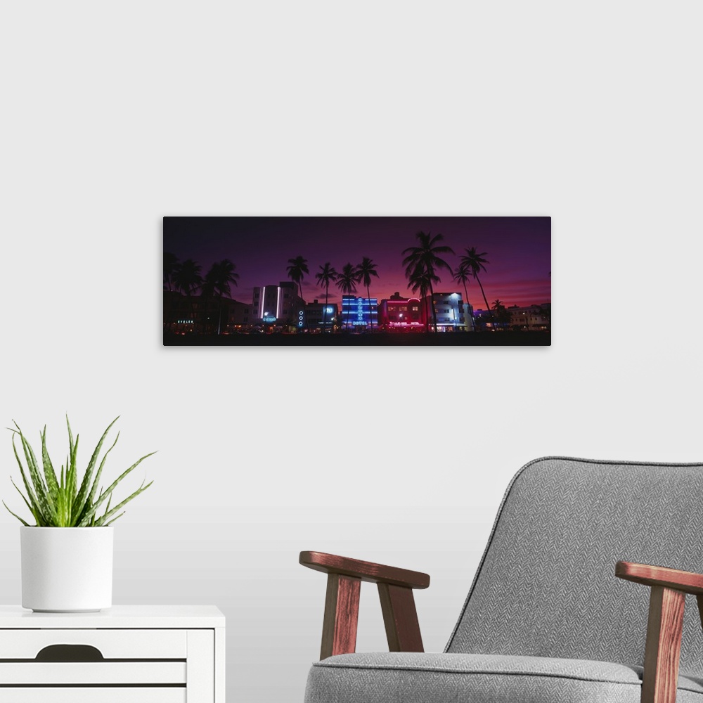 A modern room featuring A panoramic canvas of neon lights on art deco buildings on the shore front lined with palm trees.