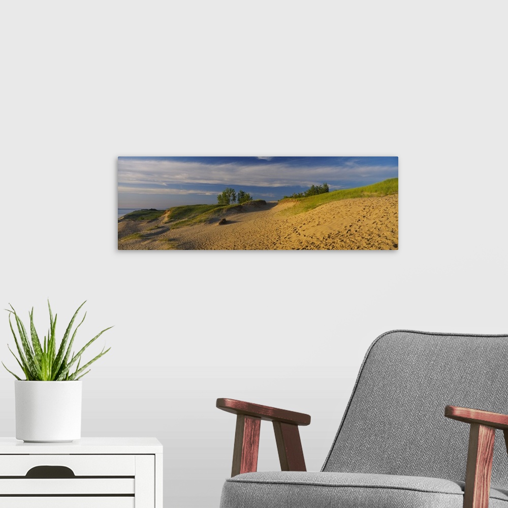 A modern room featuring Sloping sand dunes covered in beach grass and footprints under wispy clouds at sunset at the edge...