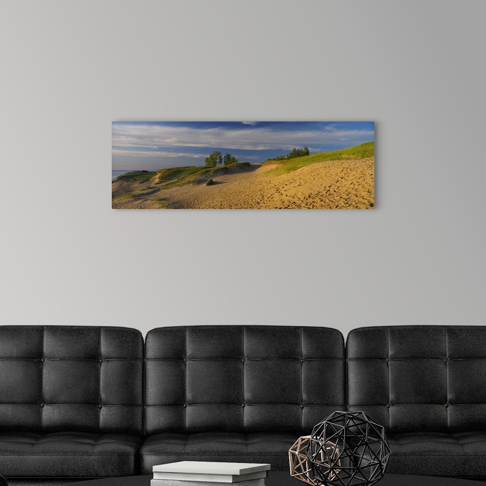 A modern room featuring Sloping sand dunes covered in beach grass and footprints under wispy clouds at sunset at the edge...