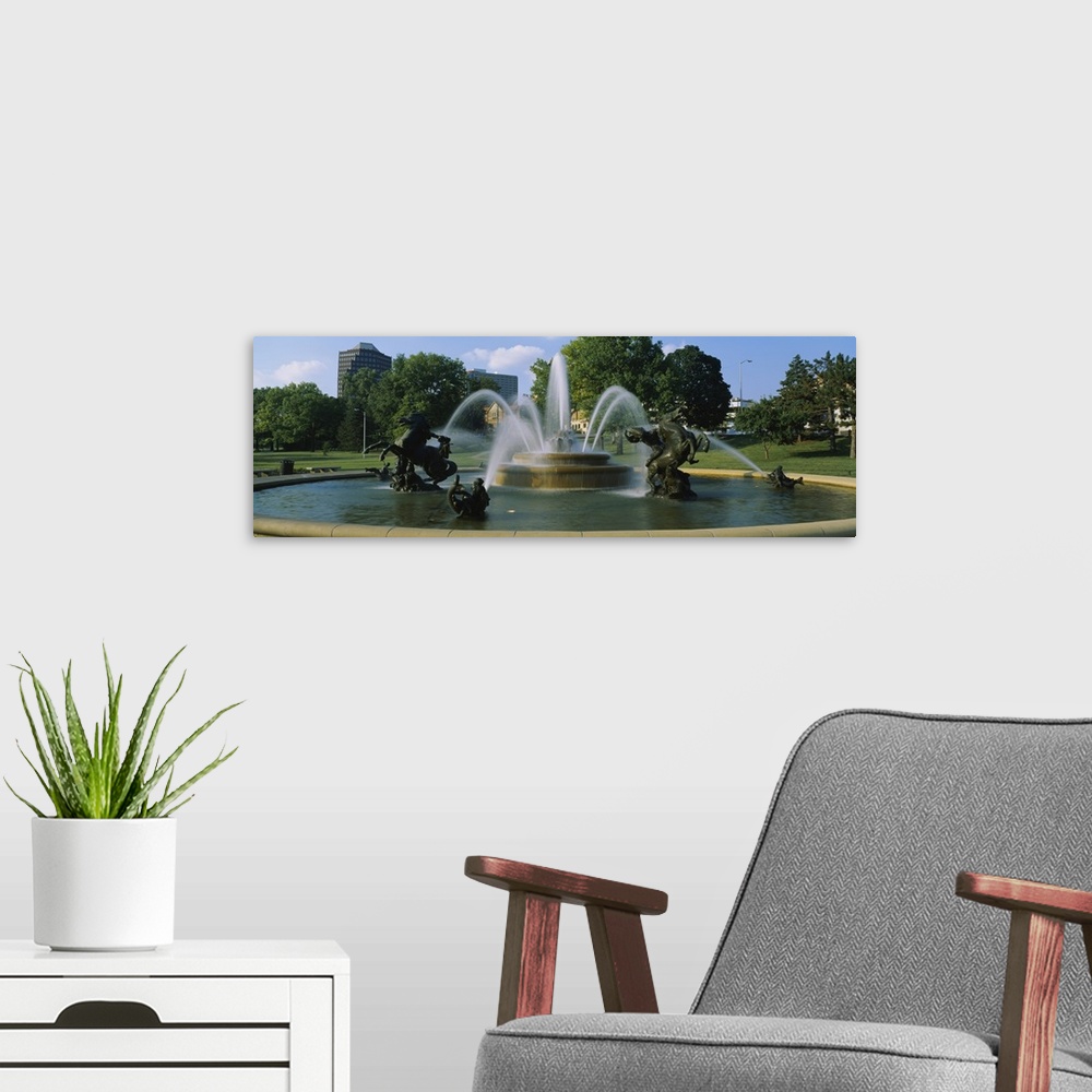 A modern room featuring A large fountain with various statues in it is photographed in panoramic view with a park shown i...