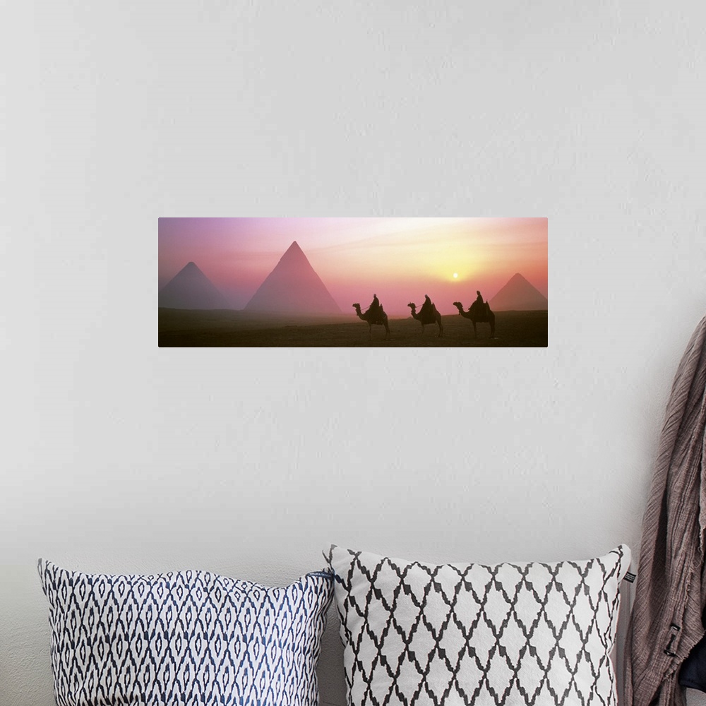 A bohemian room featuring Panoramic photograph of three camel riders at dusk with pyramids in the background.  The sun is s...