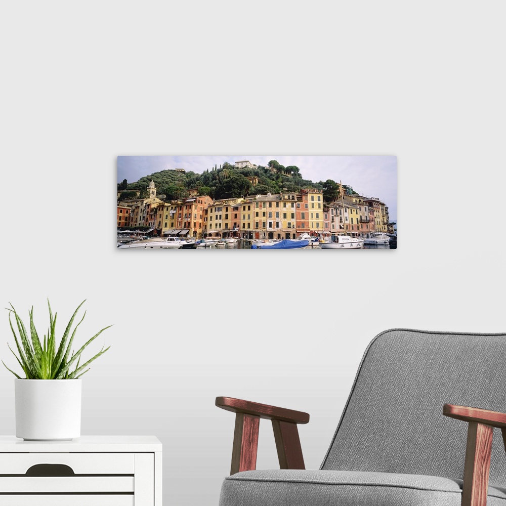 A modern room featuring Houses in Italy line a harbor that is filled with small boats. A large hill covered with trees st...