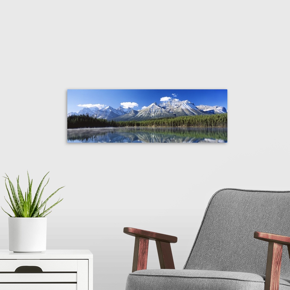 A modern room featuring Large panoramic canvas of big snowy mountains with a dense forest beneath it reflected in the water.