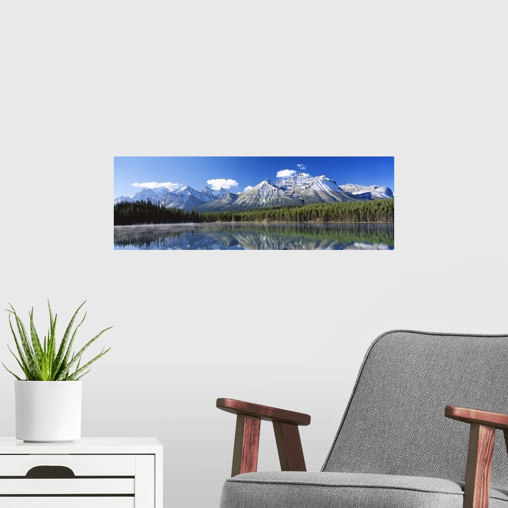 A modern room featuring Large panoramic canvas of big snowy mountains with a dense forest beneath it reflected in the water.