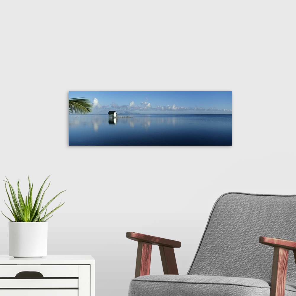 A modern room featuring Oversized landscape photograph of a small hut in the middle of a deep blue sea, a palm frond sway...