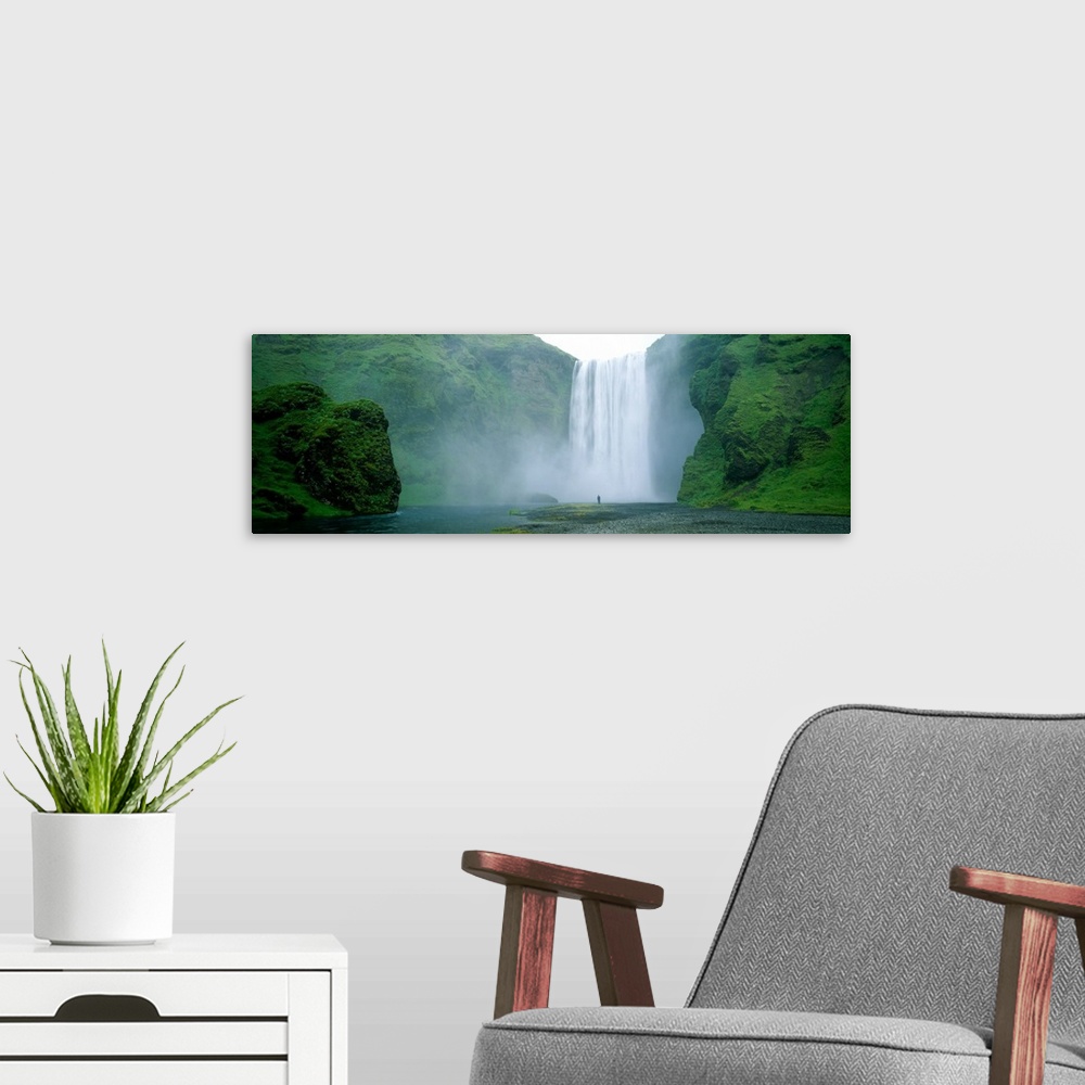 A modern room featuring Panoramic photo of a wide waterfall spilling over a cliff into the water below with a man standin...