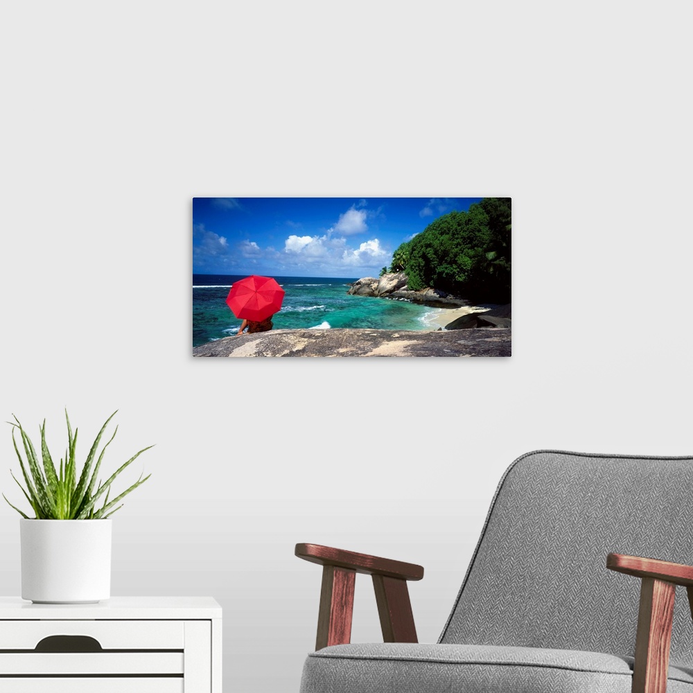 A modern room featuring A woman holding a red umbrella sits on a large rock and looks out over the ocean with the coast j...