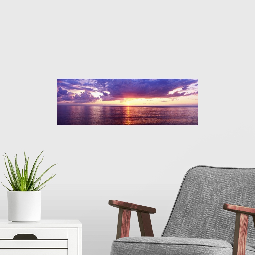 A modern room featuring Sun setting over the Great Lake, bordered by puffy clouds and the calm waters, casting its glowin...