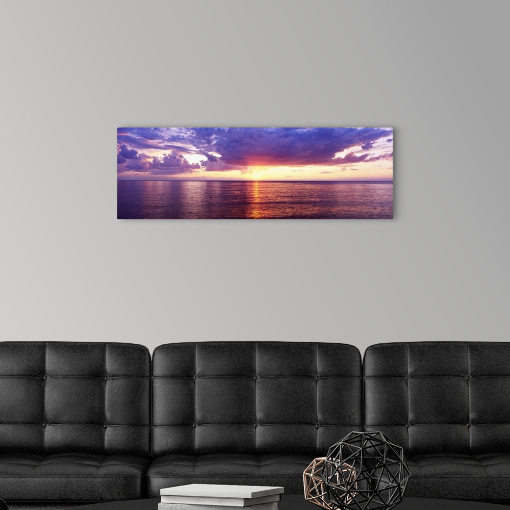 A modern room featuring Sun setting over the Great Lake, bordered by puffy clouds and the calm waters, casting its glowin...