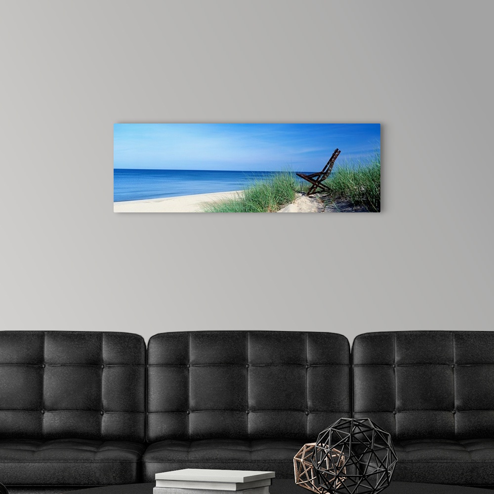 A modern room featuring This wall art is a panoramic landscape photograph of a sandy beach with a chair in the dunes over...