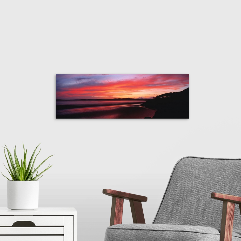 A modern room featuring A large panoramic photograph taken of a sunset over a bay in the UK. A hill top is silhouetted to...