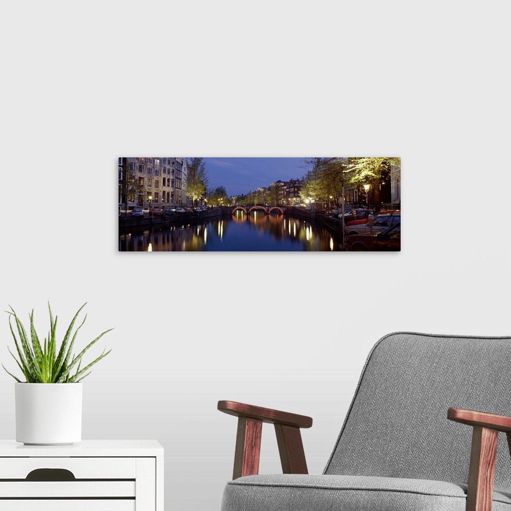 A modern room featuring A wide angle view of a canal in Amsterdam with buildings and street lights illuminated on either ...