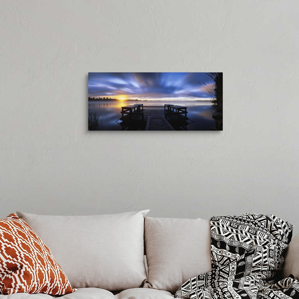 A bohemian room featuring Panoramic photograph f wooden dock at sunset with forest in distance under a cloudy sky.