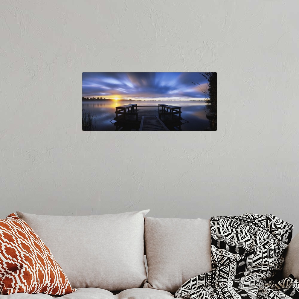 A bohemian room featuring Panoramic photograph f wooden dock at sunset with forest in distance under a cloudy sky.