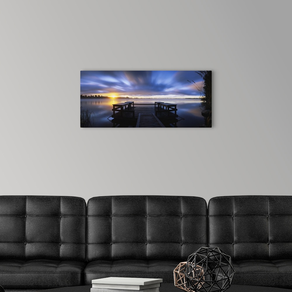 A modern room featuring Panoramic photograph f wooden dock at sunset with forest in distance under a cloudy sky.