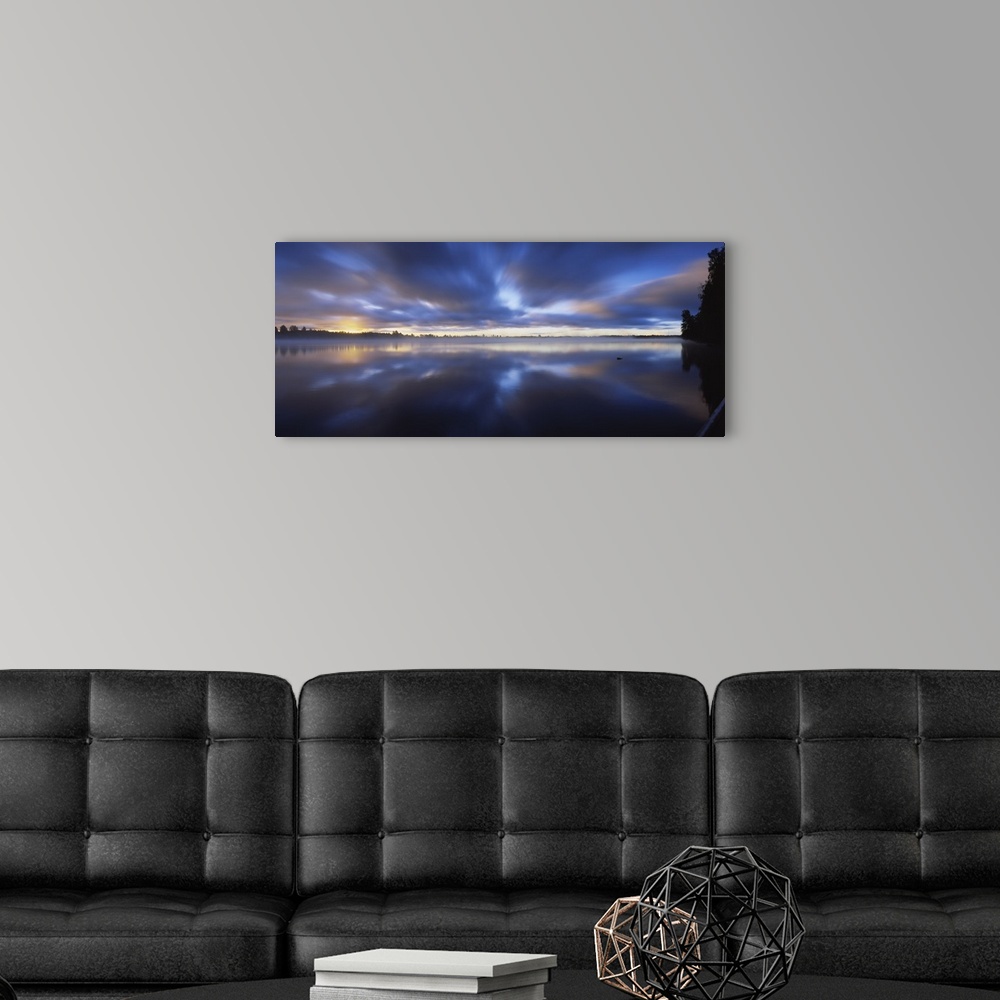 A modern room featuring Panoramic photograph of huge body of water with trees in the distance at sunset.  The sky is clou...