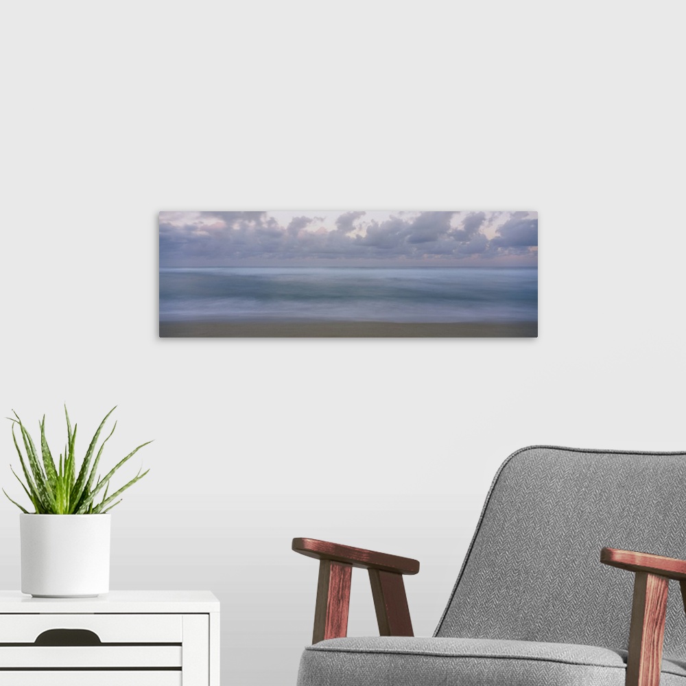 A modern room featuring This is a panoramic photograph of a waves gently washing on the sandy shore with a cloudy sky ove...