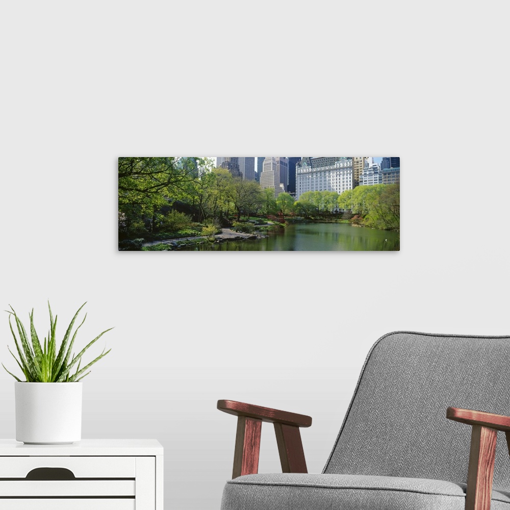 A modern room featuring A large pond in Central Park is photographed with thick foliage lining the left side and building...