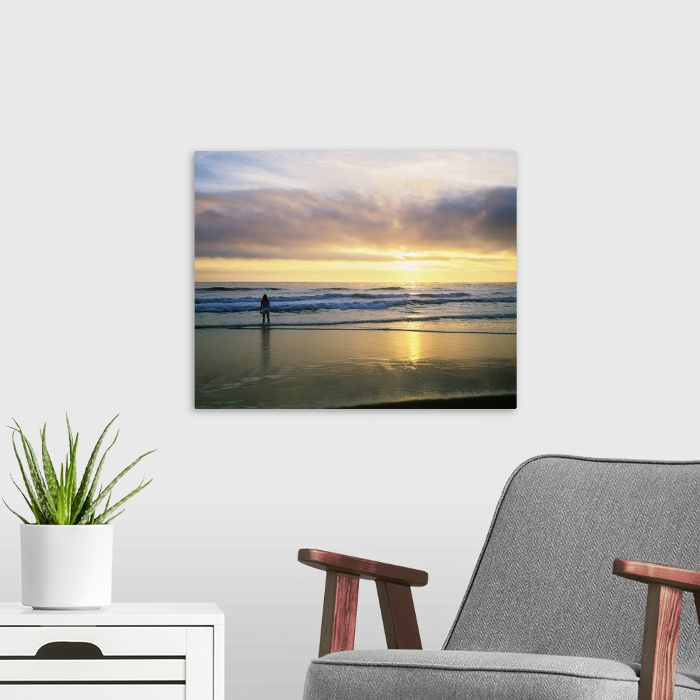 A modern room featuring In this landscape photograph a lone figure stands in the waves on the shore watching the sunset o...