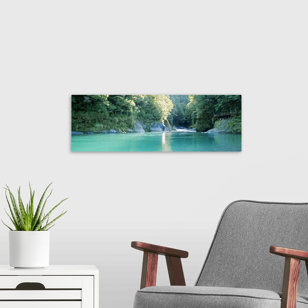 A modern room featuring Panoramic image of a river with clear water flowing through a dense forest with big rocks as clif...