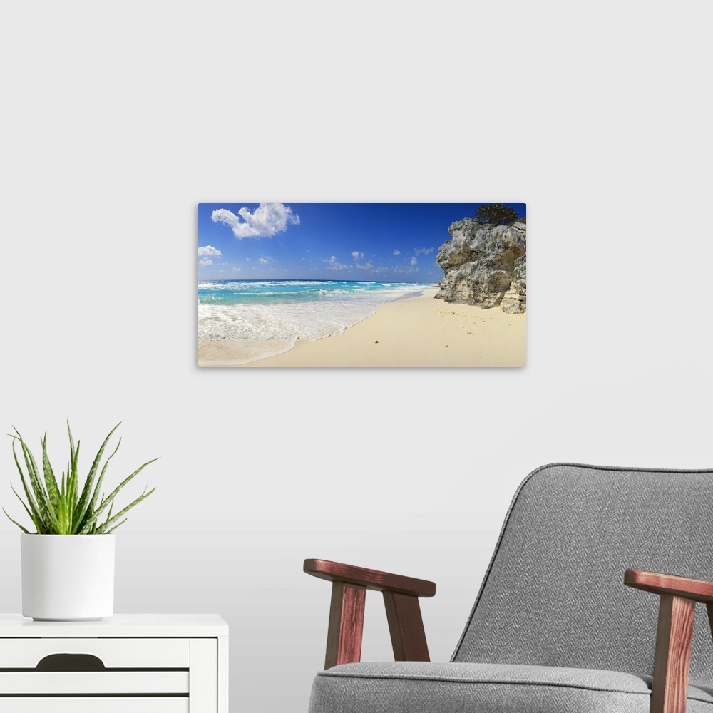 A modern room featuring Panoramic photograph of beach with huge rock structure with tree on top.  Waves and sea foam and ...