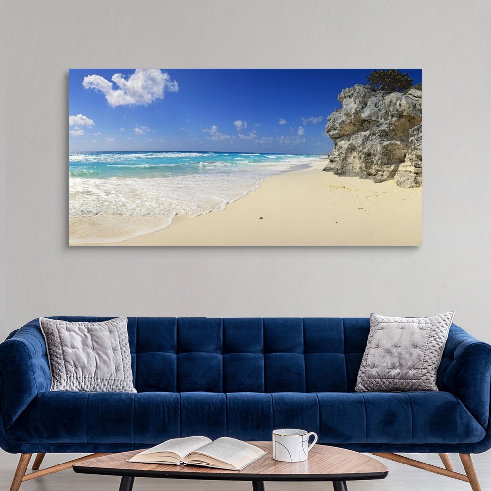 A modern room featuring Panoramic photograph of beach with huge rock structure with tree on top.  Waves and sea foam and ...