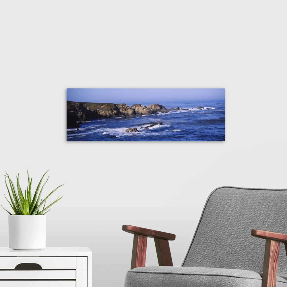 A modern room featuring Wide angle photograph of the bright blue waters of Big Sur lapping around large rock formations n...