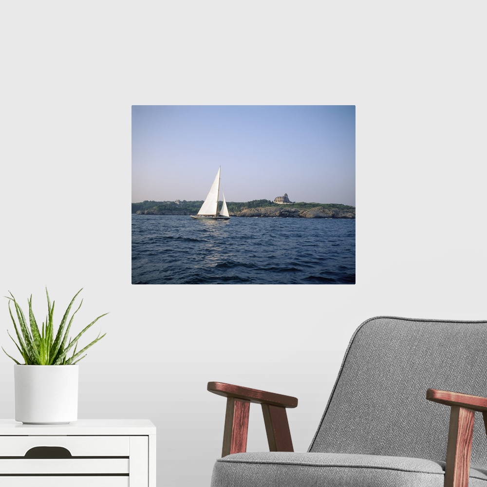 A modern room featuring A single sail boat drifts in the ocean. A large home is pictured on land behind the boat and rock...
