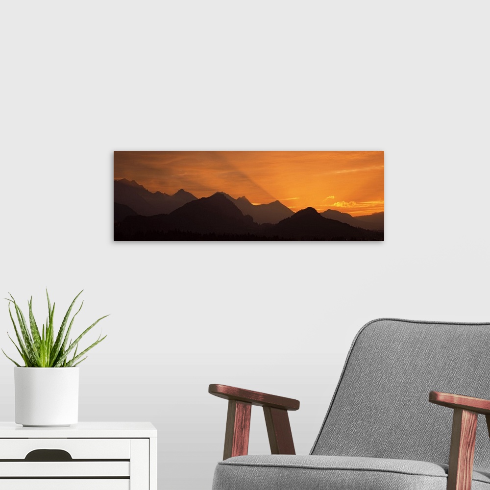 A modern room featuring Wide angle photograph of the sun setting behind the silhouetted European Alps, beneath a golden s...