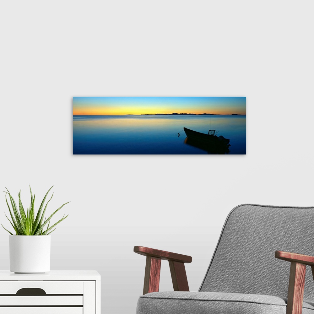 A modern room featuring Lonely boat resting on the calm ocean as the fading sun emits a golden glow on the horizon.