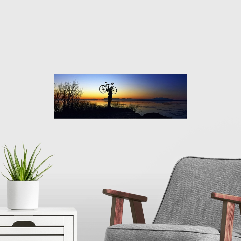 A modern room featuring Panoramic photograph of man holding bike in air at shoreline at dusk with mountains in the distance.