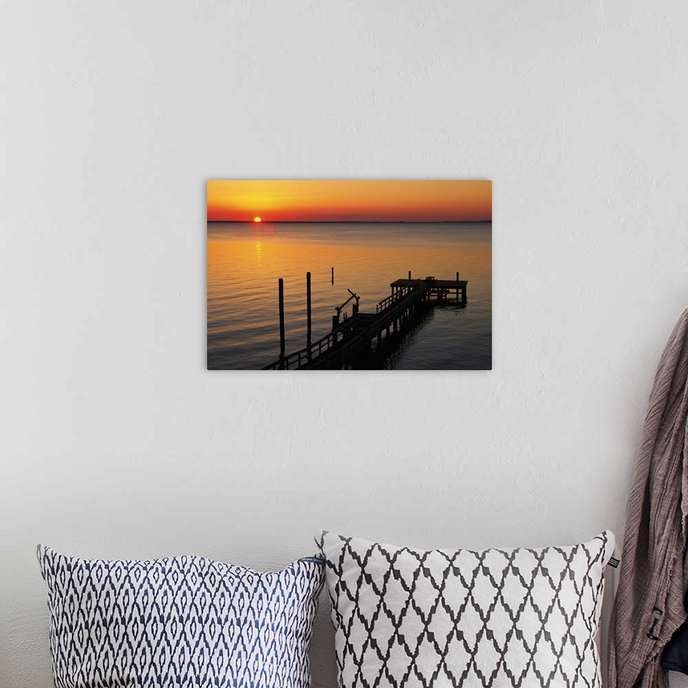 A bohemian room featuring A long pier is photographed reaching out into the ocean with the sun setting just on the horizon.