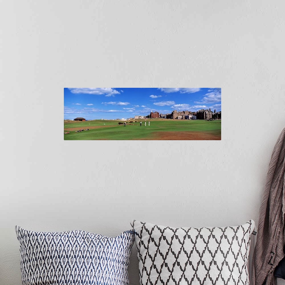 A bohemian room featuring A group of people tee off on a golf course in the UK. Buildings on the course line the right side...