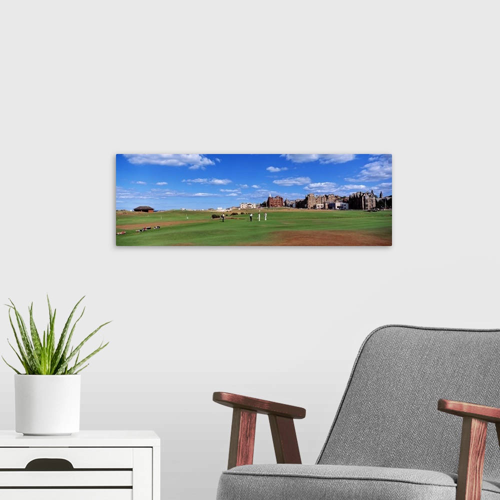 A modern room featuring A group of people tee off on a golf course in the UK. Buildings on the course line the right side...