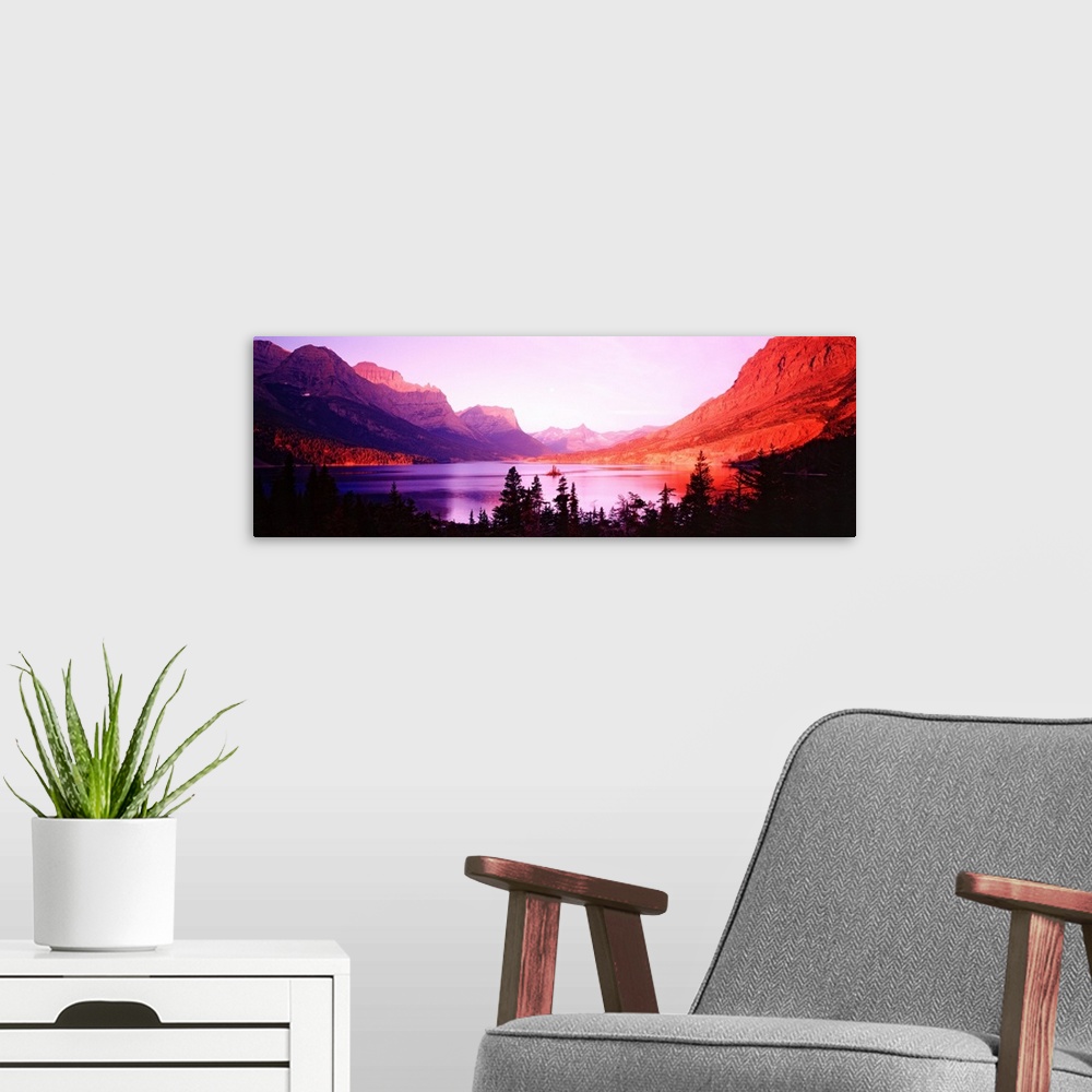 A modern room featuring Mountains and trees surrounded a  lake that reflects the colors of the setting sun on its surface...
