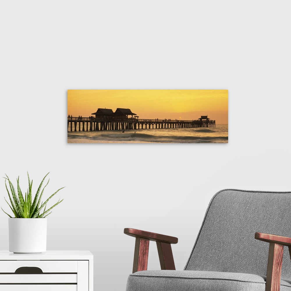 A modern room featuring A silhouetted wooden wharf with small buildings stretches out into the ocean at sunset. A couple ...