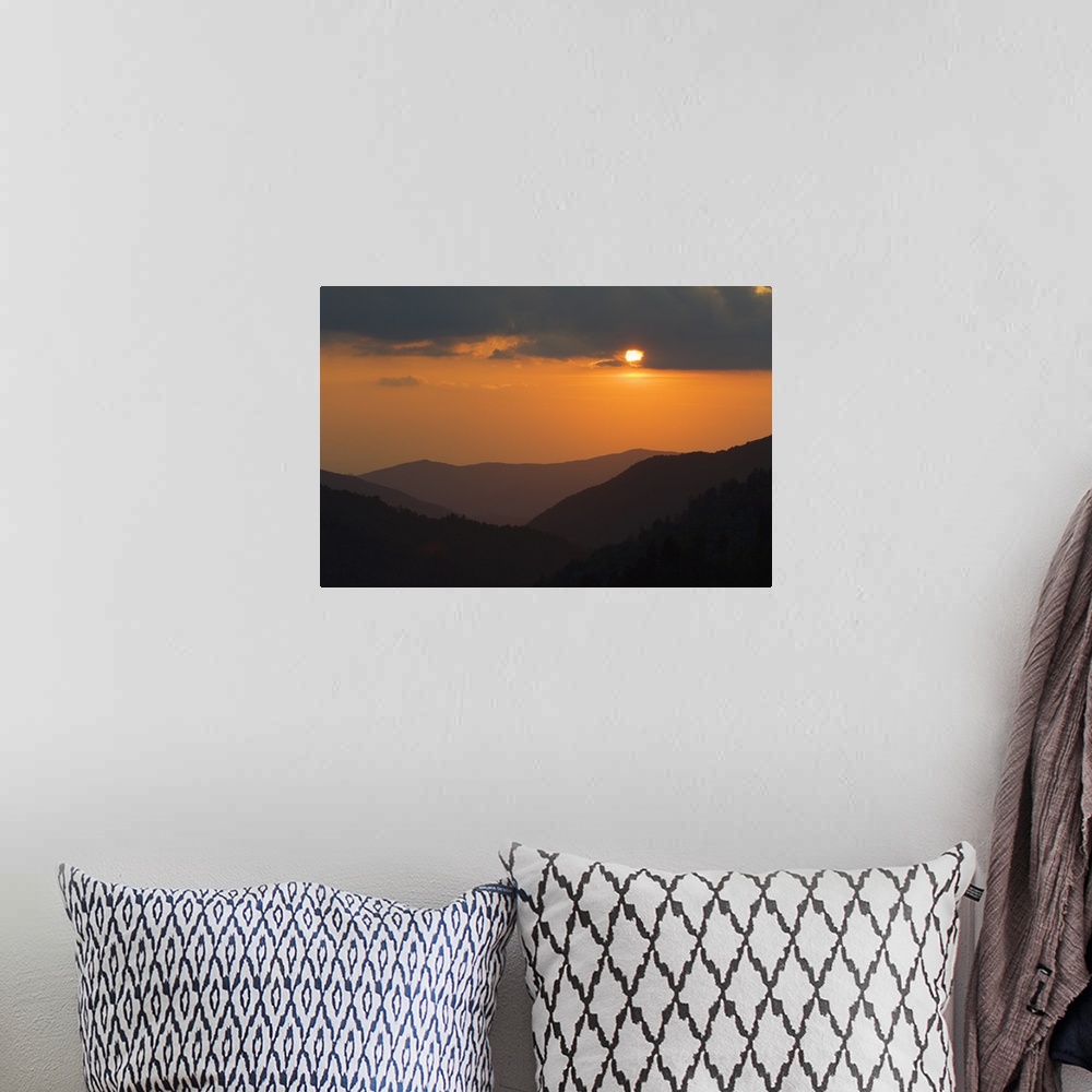 A bohemian room featuring An oversized piece of artwork that is a photograph of the sun setting behind dark clouds with tre...