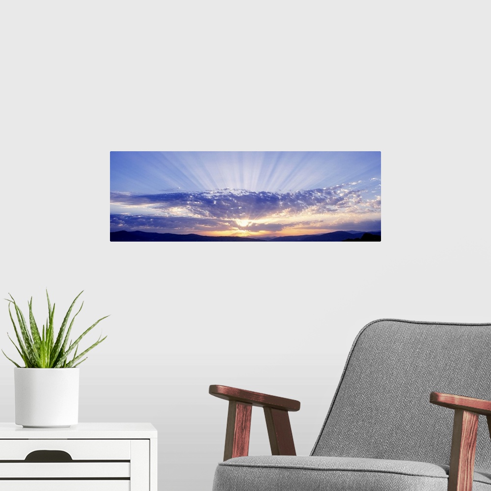 A modern room featuring This panoramic wall art captures as a photograph radiant sun beams radiate from behind clouds ove...