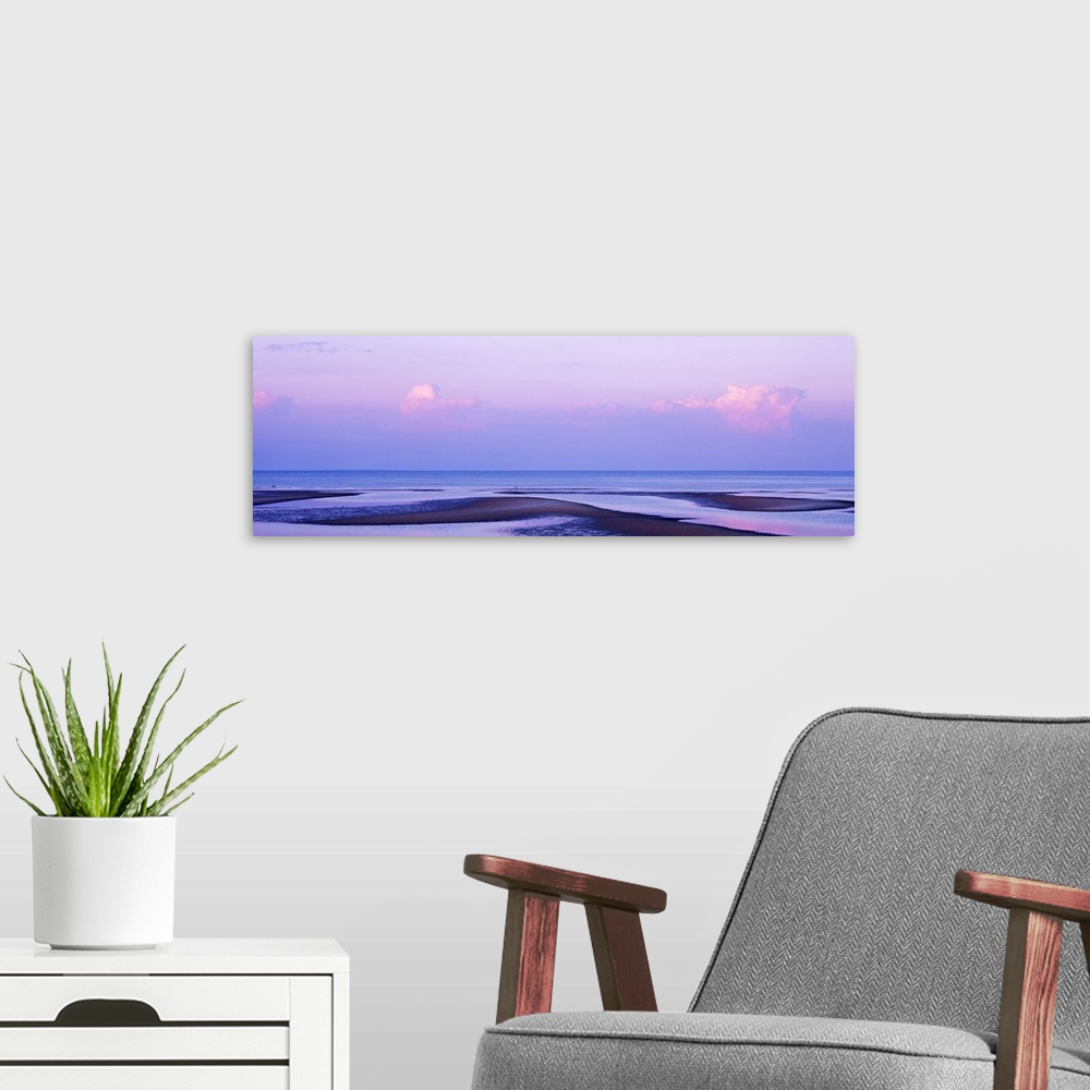 A modern room featuring An elongated view of the ocean with large water puddles on the beach and the sky has pink tones f...