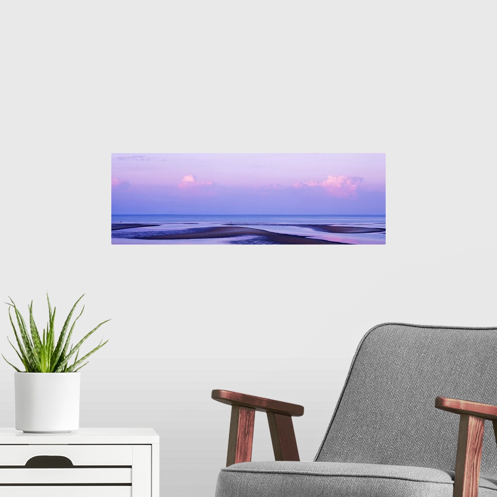 A modern room featuring An elongated view of the ocean with large water puddles on the beach and the sky has pink tones f...