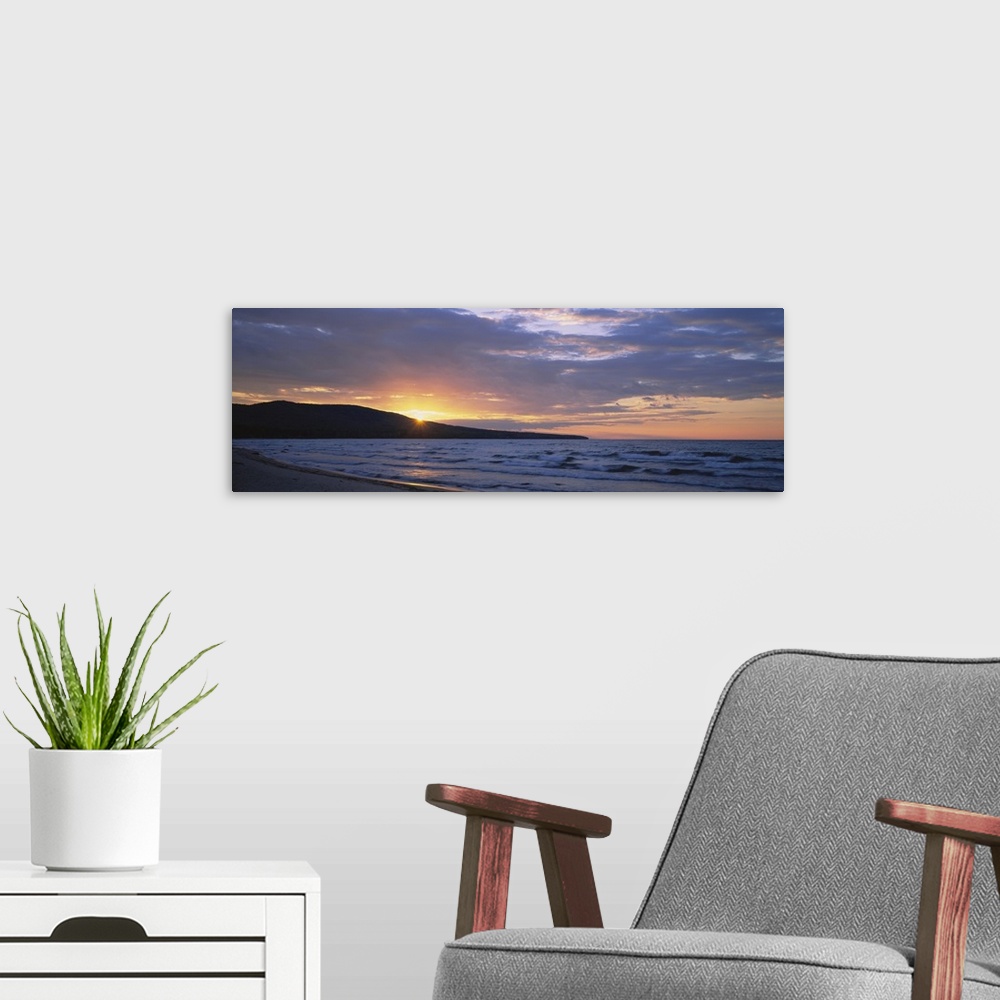 A modern room featuring Panoramic canvas photo of waves crashing in the ocean and a setting sun peeking through a rolling...
