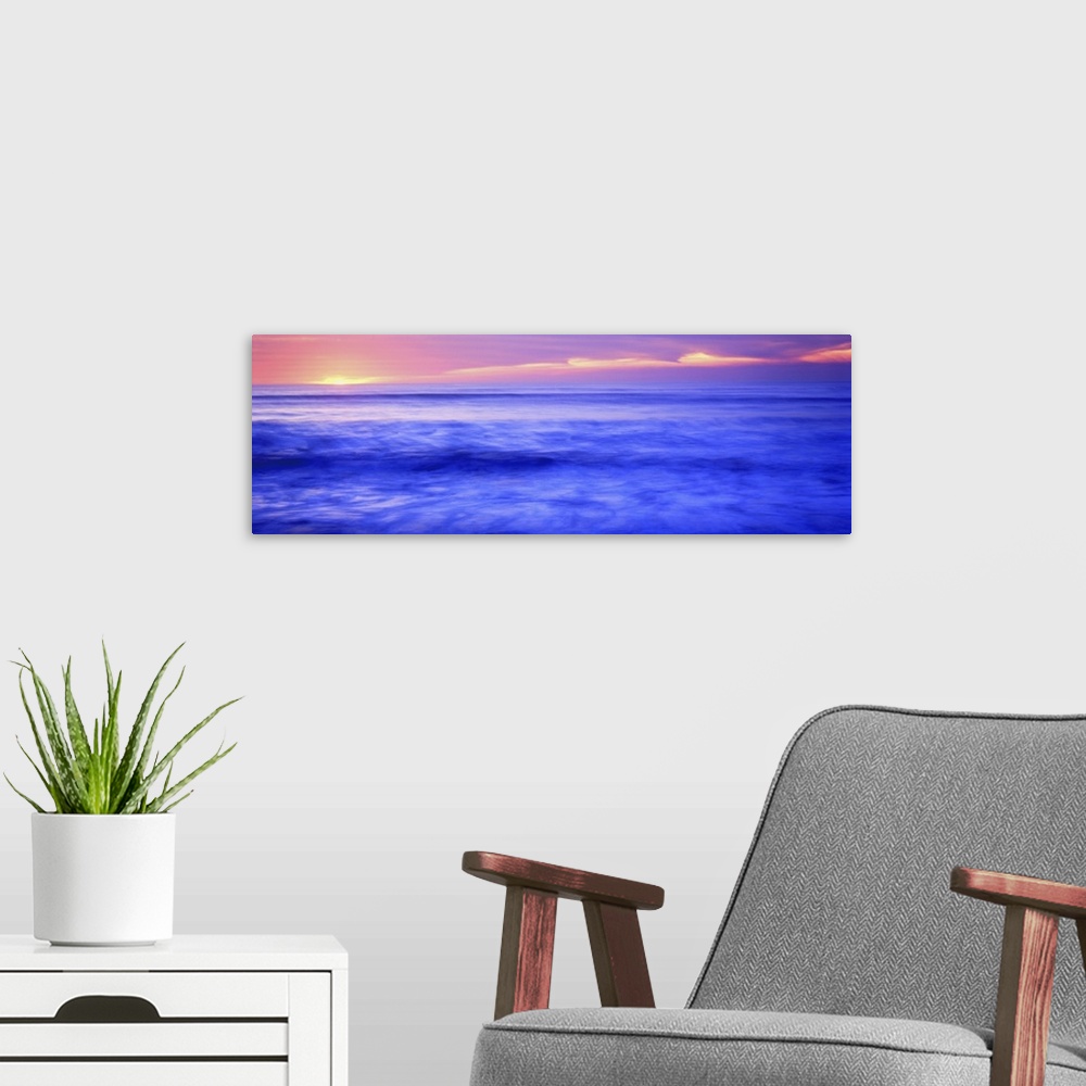 A modern room featuring Panoramic photograph shows the sun setting over the horizon and sparkling over a coast of the Wes...