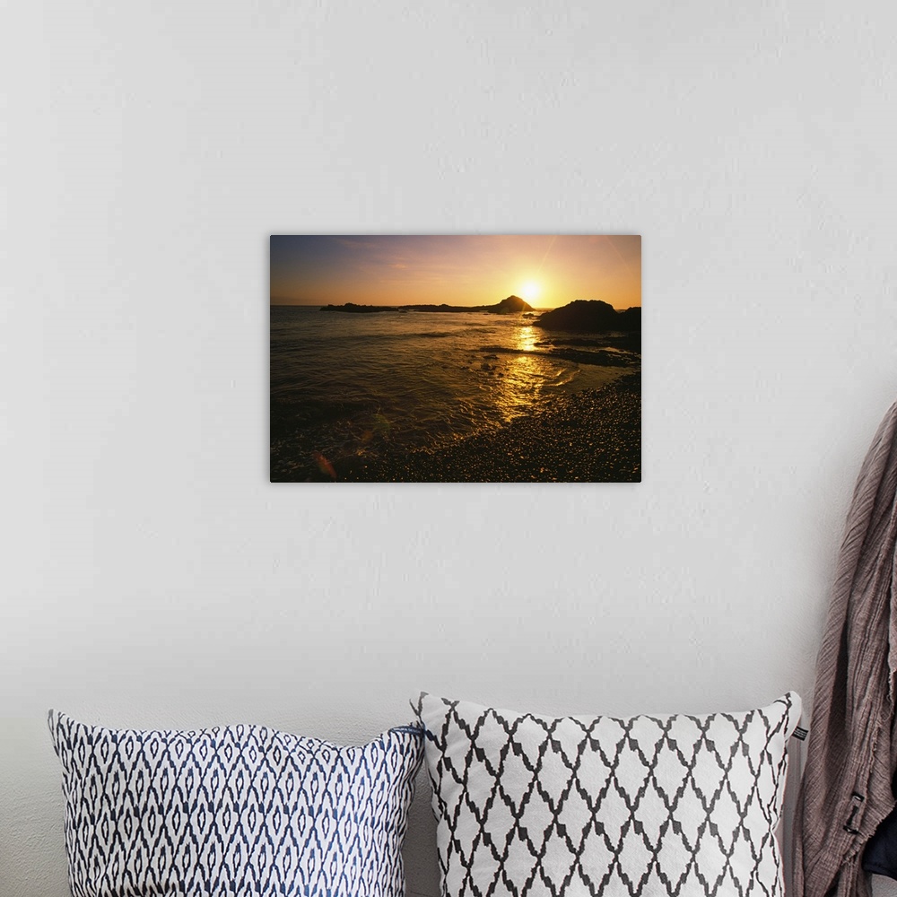 A bohemian room featuring Large, landscape photograph of the sun setting over large rocks in the water, near the shore of t...