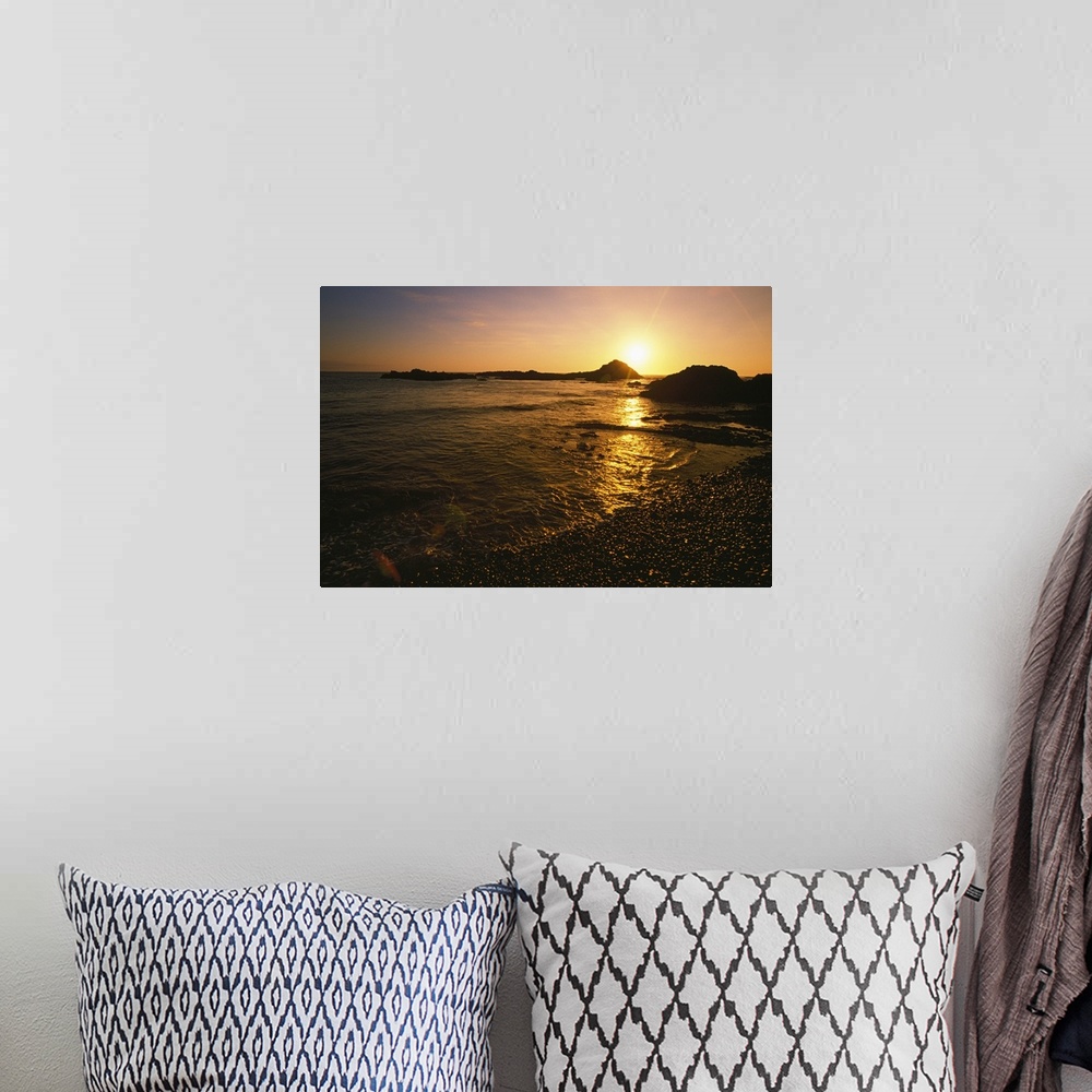A bohemian room featuring Large, landscape photograph of the sun setting over large rocks in the water, near the shore of t...