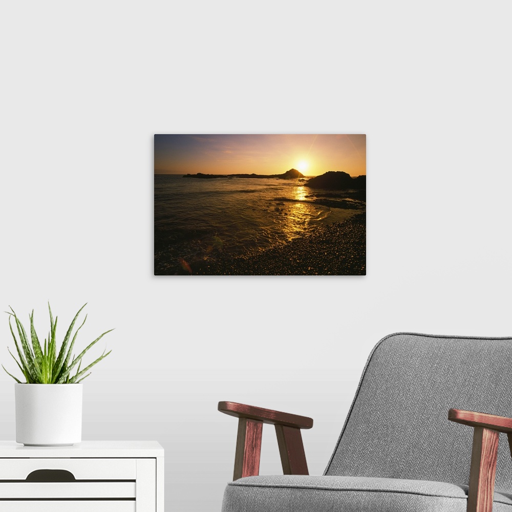A modern room featuring Large, landscape photograph of the sun setting over large rocks in the water, near the shore of t...