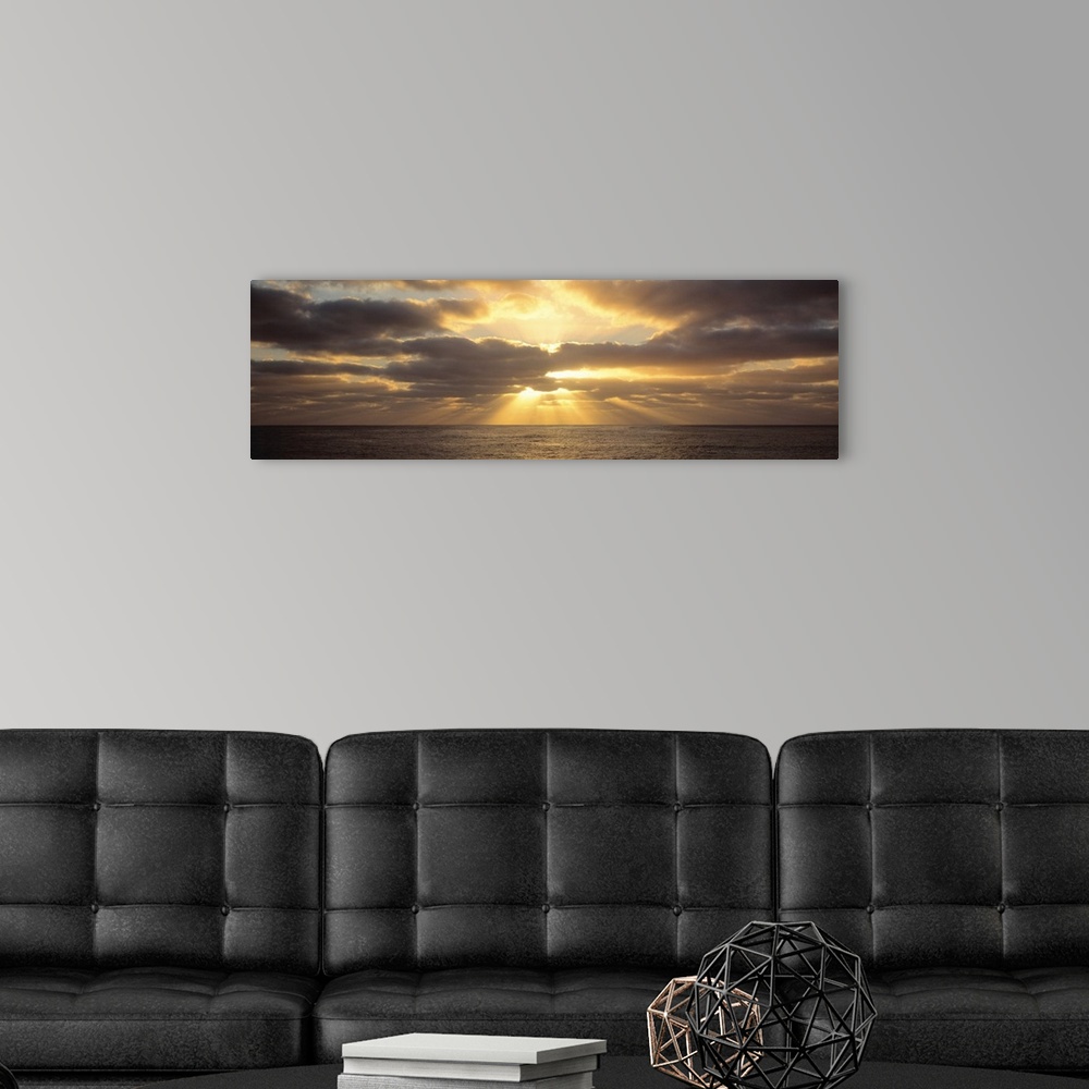 A modern room featuring Panoramic photograph on a big canvas of a sunset beaming through large clouds, over the water in ...
