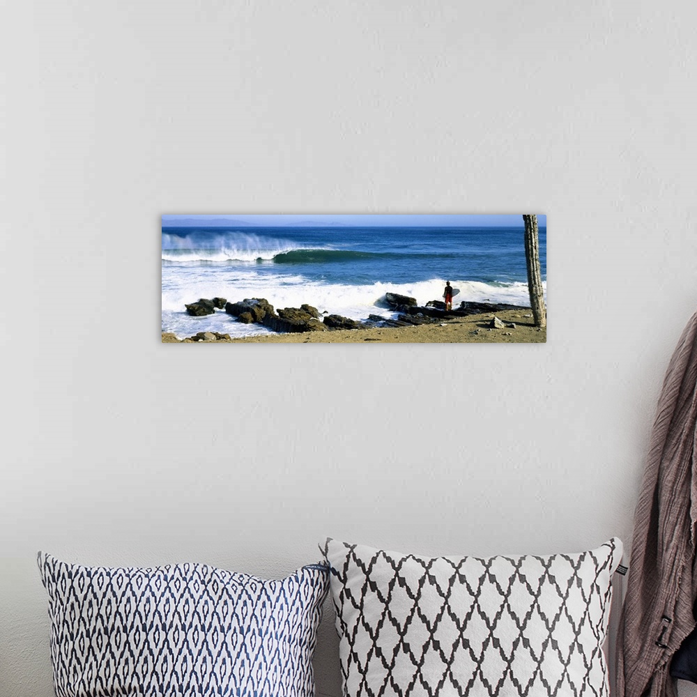 A bohemian room featuring A single figure stands amongst rocks watching waves break on the shore in this panoramic photograph.